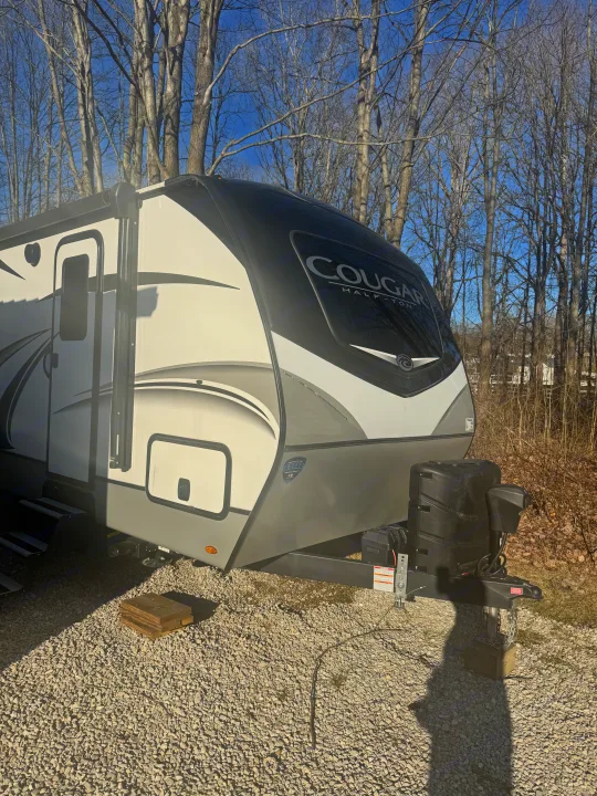 A white and black Cougar RV rental is parked on a gravel campsite. Bare trees stand against a blue sky, ideal for camping.. Keystone RV Cougar 2022