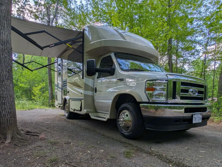 Class C RV rental with awning extended, parked on a gravel path in a wooded Virginia campsite.. Gulf Stream B Touring Cruiser 2021