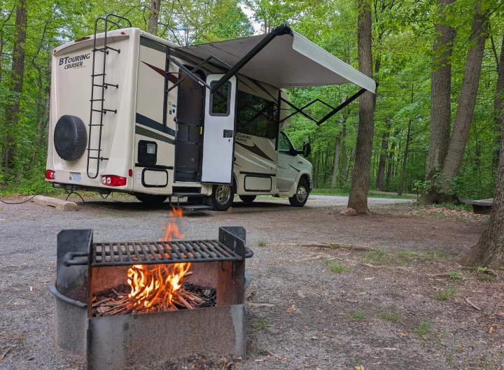An RV rental parked in a tranquil campsite, featuring an extended awning and a warm campfire in the foreground.. Gulf Stream B Touring Cruiser 2021