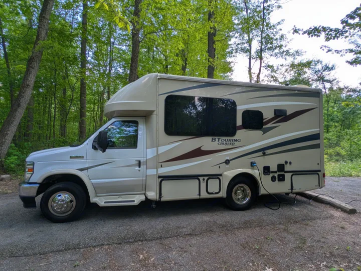 Tan B Touring Cruiser motorhome RV rental parked in a wooded campsite with tall green trees.. Gulf Stream B Touring Cruiser 2021