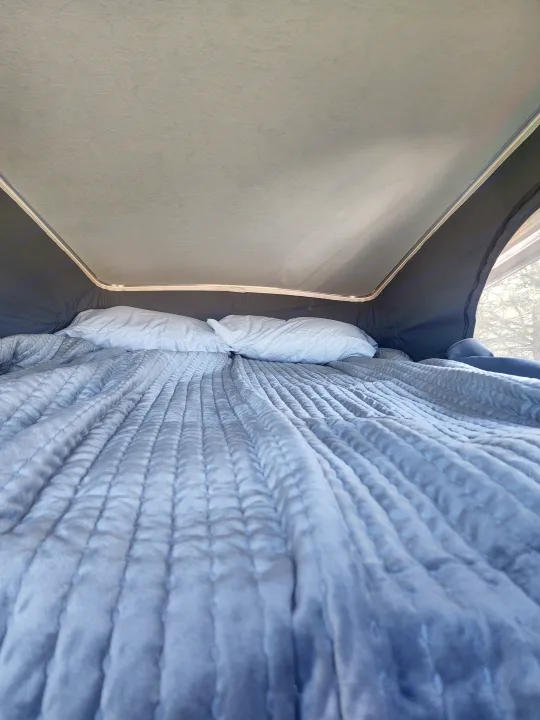 Cozy pop-top RV rental bed. A comfortable quilted grey blanket and two white pillows are ready for rest. Trees outside a mesh window.. Winnebago Solis 2022