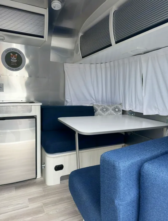 Modern RV rental interior: Dinette with blue benches, white table, and stainless fridge in a travel trailer.. Airstream Bambi 2022