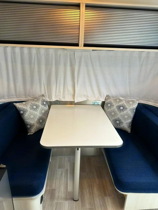 An RV rental interior showing a dinette with blue benches, a white table, and privacy blinds.. Airstream Bambi 2022