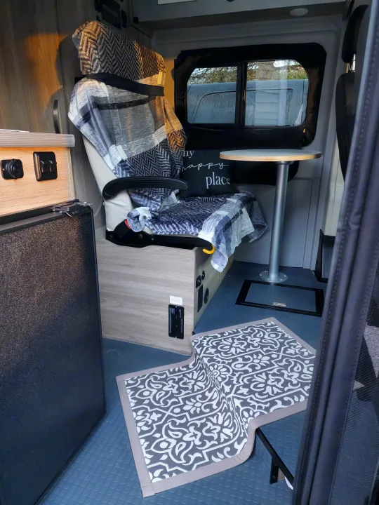Inside a cozy RV rental: a comfy patterned chair with blankets, a small table, and a fridge on a textured floor.. Winnebago Solis 2022