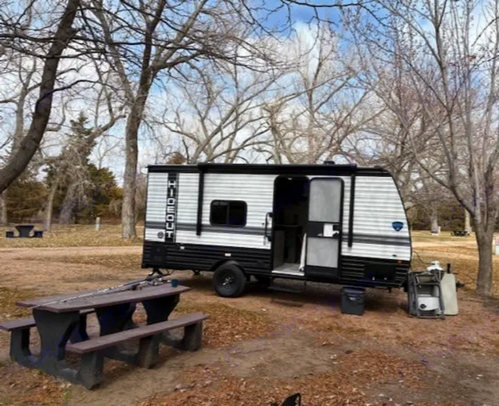 An RV rental, a white travel trailer, is set up at a peaceful campsite with a picnic table and trees.. Keystone RV Hideout Sport 2024