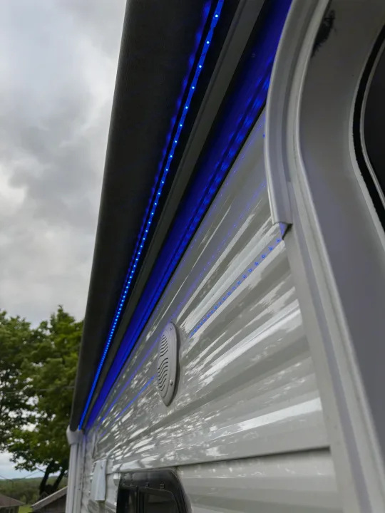 Vibrant blue LED lights under the awning of this RV rental, illuminating the white ribbed side and outdoor speaker.. Forest River Grand River Comfortline 2021