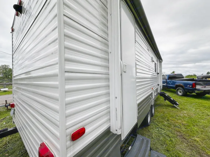 Side view of a white and grey RV rental travel trailer parked on grass under a cloudy sky. A blue truck is in the background.. Forest River Grand River Comfortline 2021