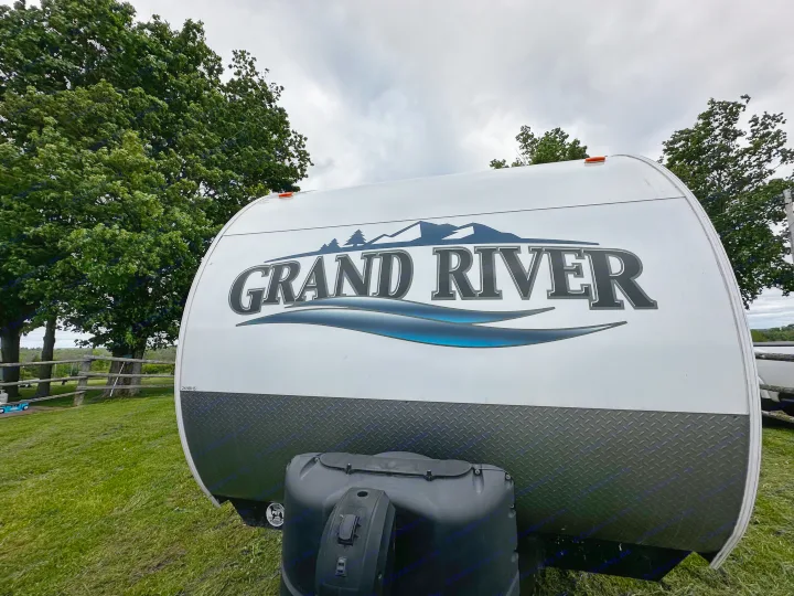 Front of a white Grand River rv rental with a mountain and river logo, parked in a grassy camp setting.. Forest River Grand River Comfortline 2021