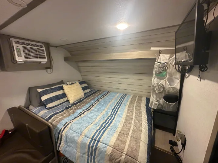 Cozy RV rental bedroom with striped bed, wall-mounted AC, TV, and handy storage, ideal for compact living on the road.. Coleman Lantern 17B 2024