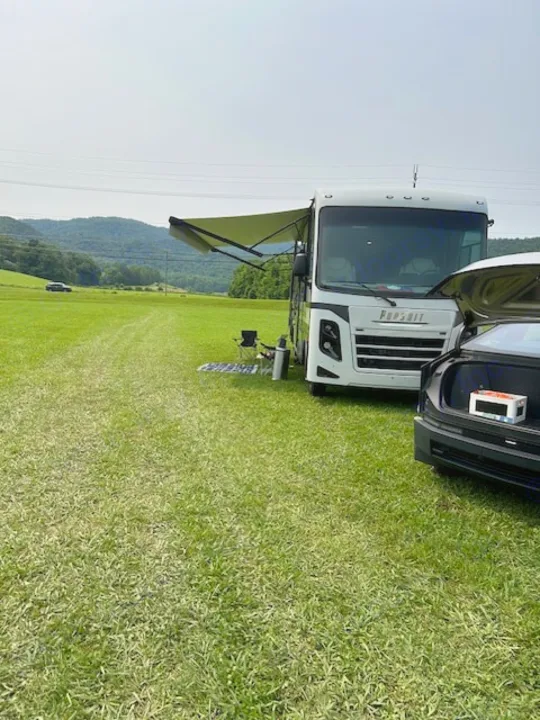 A white rv rental with its awning out, set up in a wide green field next to a car with its trunk open for a relaxing outdoor trip.. Coachmen Pursuit 31bh 2023