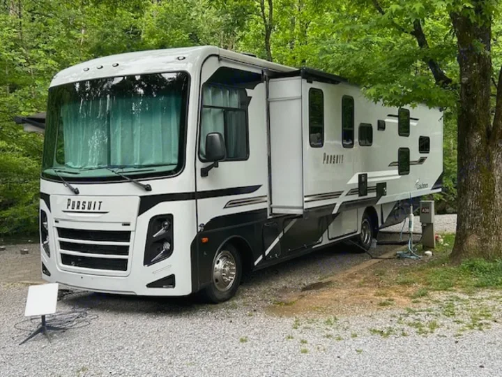White Coachmen Pursuit RV rental at a scenic gravel campsite. This spacious motorhome offers comfortable camping with Starlink internet.. Coachmen Pursuit 31bh 2023