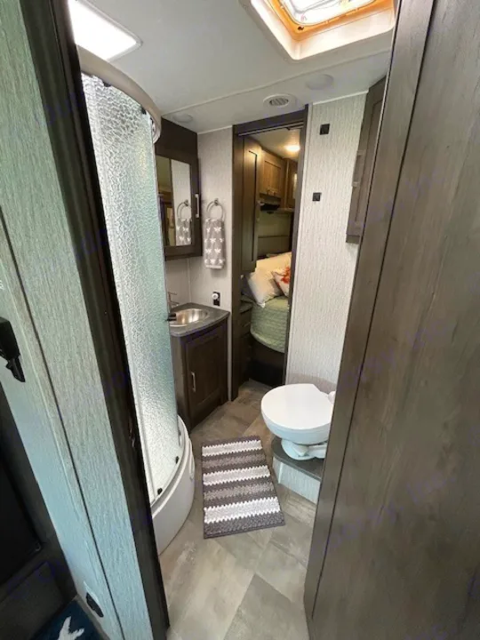 This image displays a compact RV rental bathroom with a curved shower, vanity, toilet, and a doorway leading to the bedroom.. Coachmen Pursuit 31bh 2023