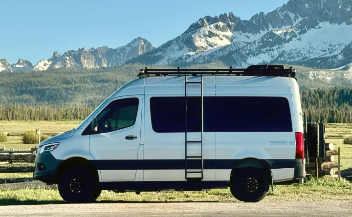 A silver RV rental camper van with roof rack and ladder, parked against a stunning backdrop of snow-capped mountains.. Cahaba Cahaba Class B 2021