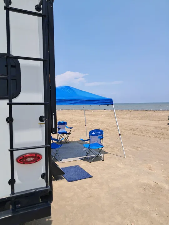 Relax by the ocean! This RV rental set up on a sandy beach features a blue canopy and chairs, offering a perfect outdoor escape.. Jayco Jay Feather 2022