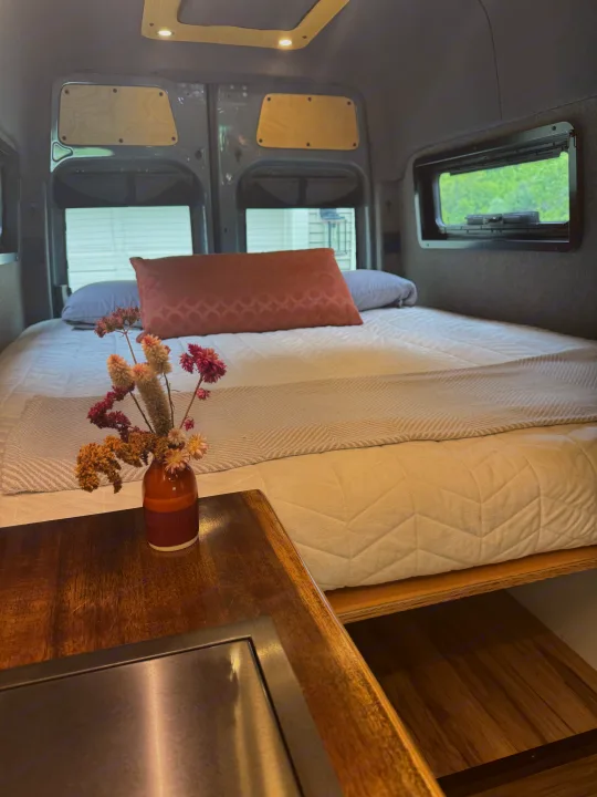 Relax in this well-appointed RV rental. A comfortable bed, warm wooden accents, and a touch of nature create a cozy travel space.. Mercedes-Benz Sprinter 2021