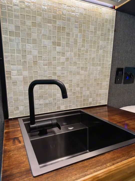 Modern RV rental kitchenette features a black faucet, dark sink, wood counter, and sleek mosaic tile backsplash for comfortable travel.. Mercedes-Benz Sprinter 2021