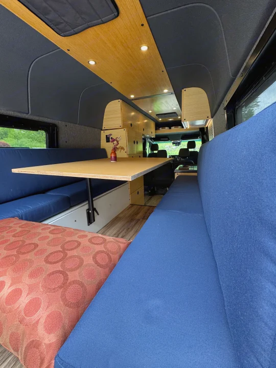 Spacious RV rental interior featuring a wooden dining table, blue upholstered benches, and custom cabinetry for comfort.. Mercedes-Benz Sprinter 2021