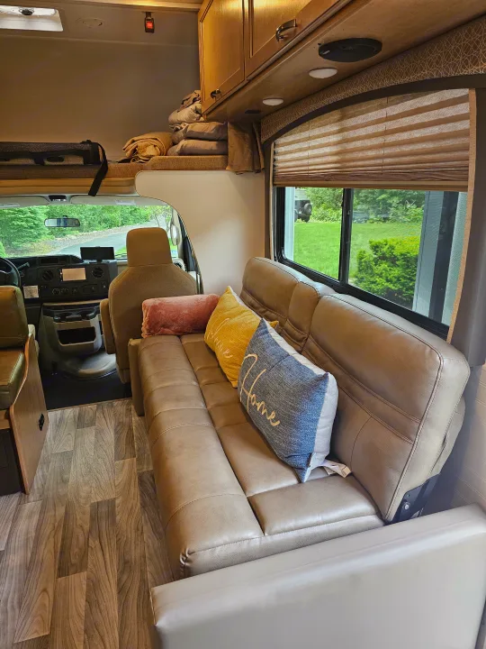 Inside a spacious RV rental: a plush sofa with colorful pillows, an overhead bunk, and front cockpit view.. Thor Four Winds 2021