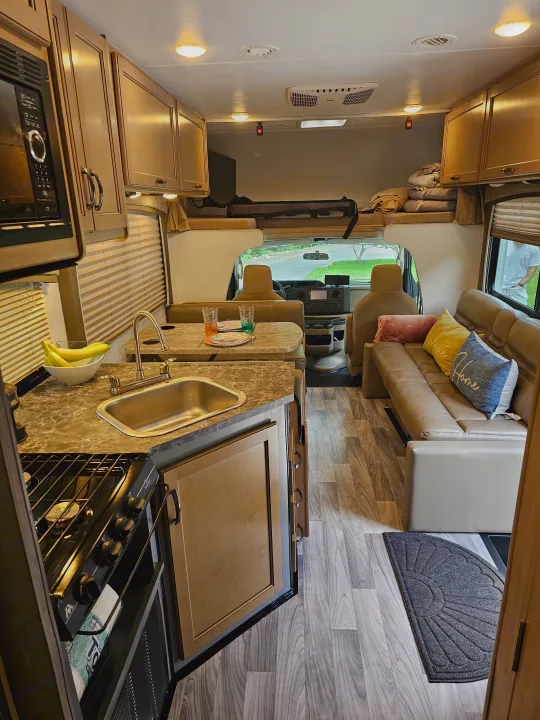 Inside a well-appointed RV rental: full kitchen with sink/stove, dining booth, plush sofa, and overhead sleeping bunk.. Thor Four Winds 2021