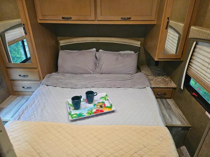 Cozy RV rental bedroom with a gray-sheeted bed, tropical tray, overhead cabinets, and side storage units with mirrors.. Thor Four Winds 2021