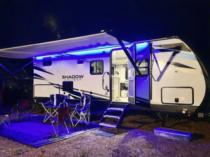 Nighttime rv rental setup: a white camper with blue lights under its extended awning, featuring outdoor chairs and a rug.. Cruiser RV Shadow Cruiser 2019