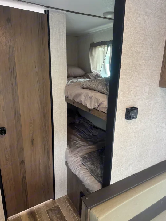 Inside an RV rental: two compact bunk beds with soft bedding, a nearby wooden door, and wall-mounted climate control.. Gulf Stream Ameri-Lite Super Lite 2022