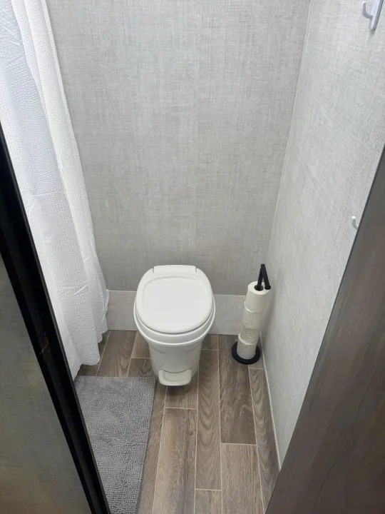 Compact RV rental bathroom: white toilet, shower curtain, wood-look floor, and textured grey walls.. Gulf Stream Ameri-Lite Super Lite 2022