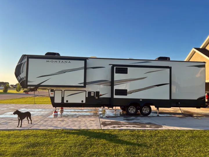 A large, white Montana High Country RV and a dark dog on a sunny, wet driveway. This type makes a great RV rental.. Keystone RV Montana High Country 2021