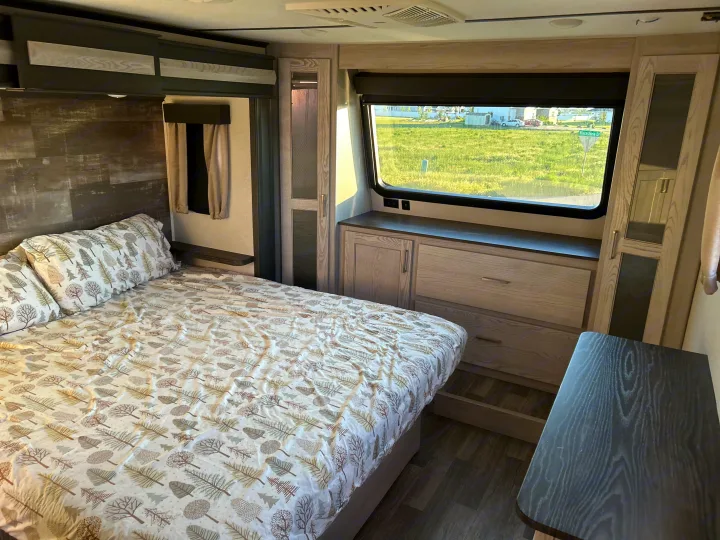 Bright RV rental bedroom featuring a bed with tree-patterned sheets, integrated storage, and a large window overlooking a field.. Keystone RV Montana High Country 2021