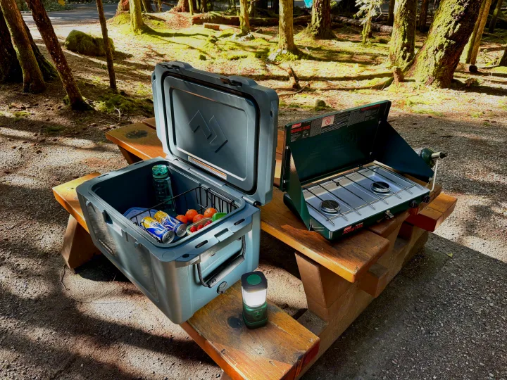Campsite gear for an RV rental trip: A gray cooler with snacks/drinks, a Coleman propane stove, and a lantern on a picnic table.. Ford Maverick 2025