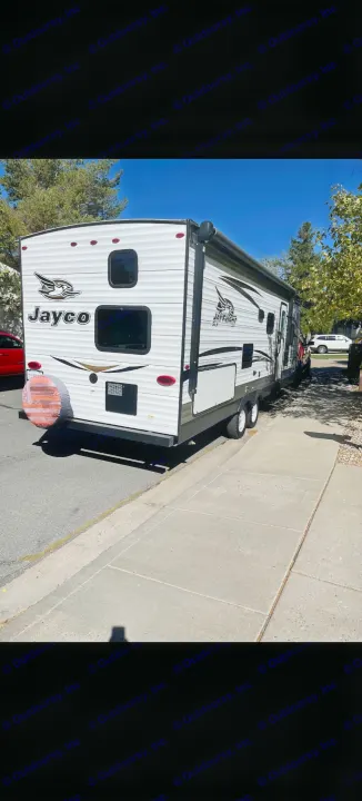 A white Jayco Jay Flight SLX travel trailer, ideal for your next RV rental, is parked on a sunlit residential street with a sidewalk.. Jayco Jay Flight 2018