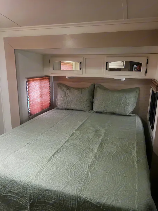 An RV rental bedroom featuring a neatly made bed with light green quilted bedding, two pillows, mirrored cabinets, and a window.. Keystone RV Outback Toy Hauler 2013