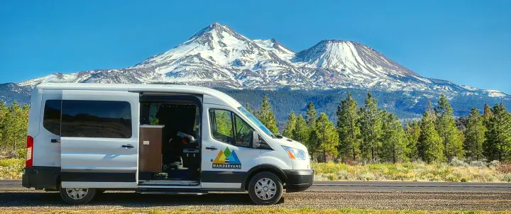 Experience scenic travels! This RV rental van is parked with its door open, showcasing snow-capped mountains and pine forests.. Ford Transit 2019