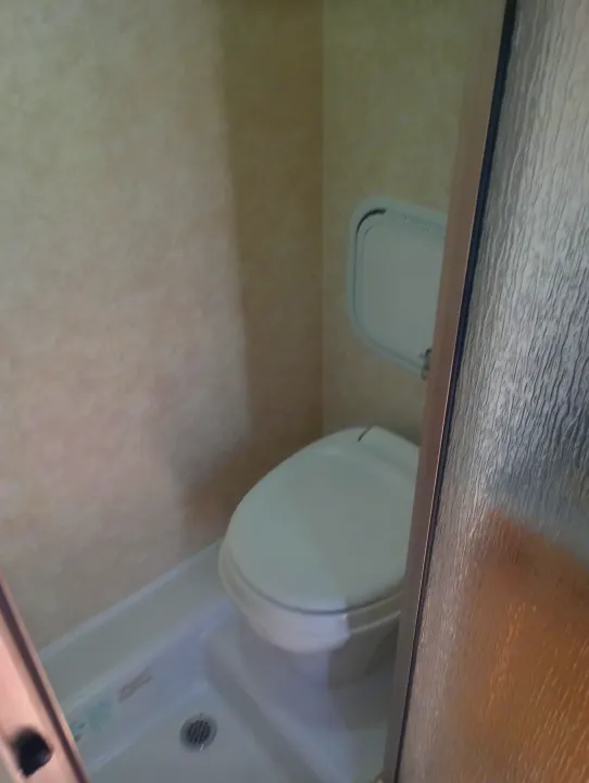 Compact RV rental bathroom featuring a toilet, shower, small wall panel, and textured glass door.. nuCamp T@B 2019