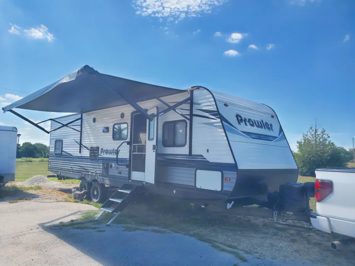 This Prowler travel trailer with extended awning and open door, perfect for an RV rental adventure under a blue sky.. Heartland RVs Prowler 2021