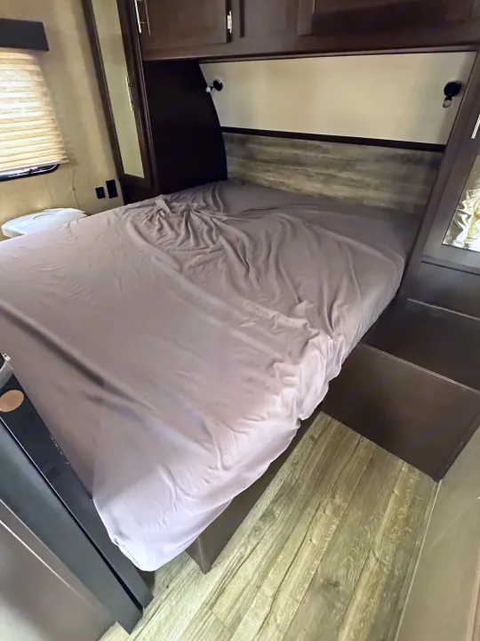 Cozy RV rental bedroom featuring a bed with rumpled sheets, overhead cabinets, and a side window with blinds.. Forest River Cherokee Grey Wolf 2017