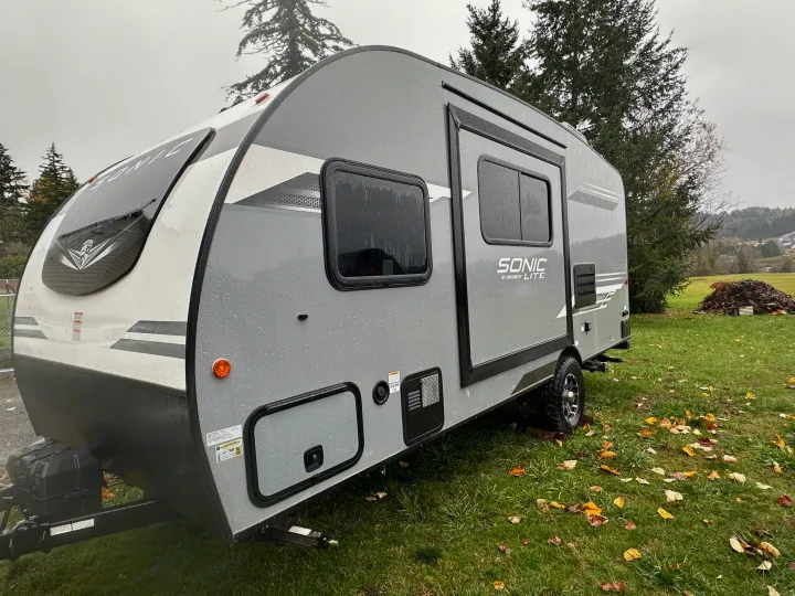 A sleek grey Sonic Lite travel trailer, ready for your next RV rental adventure, parked on a wet grassy field with trees.. Venture RV SL169VUD 2023