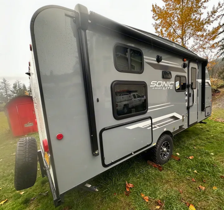 Gray Sonic travel trailer for your next RV rental trip, parked on wet grass with trees and a red shed in background.. Venture RV SL169VUD 2023
