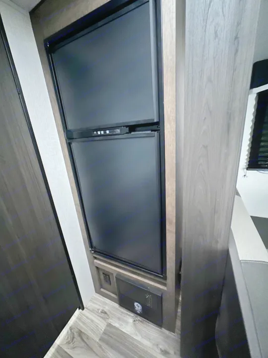 Inside this RV rental, a black refrigerator is framed by wood cabinetry, with flooring, an outlet, and seating visible.. Venture RV SL169VUD 2023
