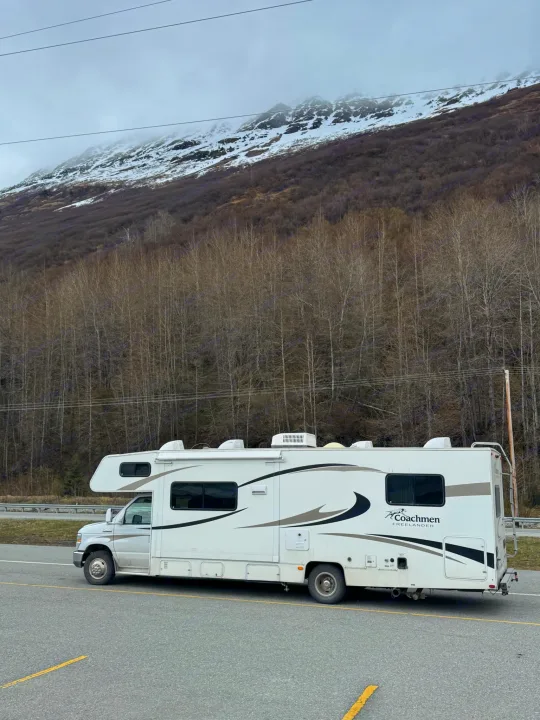 An RV rental is parked by a road, set against a snow-capped mountain and bare forest, perfect for a scenic adventure.. Coachmen Freelander 2014