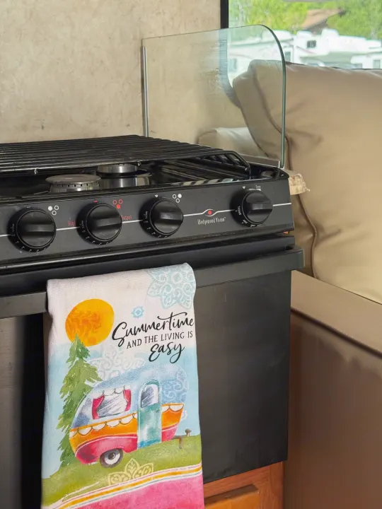 RV rental kitchen: a detailed view of a stove with controls, a glass guard, and a cheerful "Summertime" towel.. Coachmen Freelander 2014