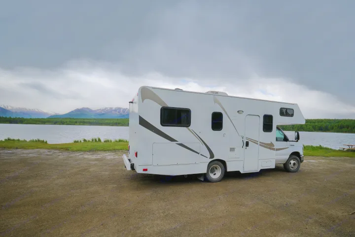 White RV rental parked on a gravel shore, overlooking a lake with forested hills and distant snowy mountains.. Thor Majestic 2018