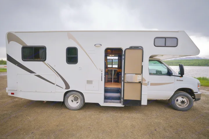 White RV rental parked by a scenic lake. Its side door is open, revealing the interior, with mountains in the distance.. Thor Majestic 2018