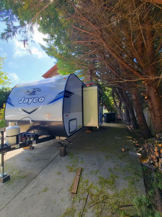 Sunny RV rental scene: Jayco travel trailer parked on concrete with slide-out extended, surrounded by tall trees and wood.. Jayco Jay Flight SLX 197MBW Baja Edition 2025