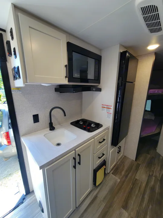 Modern RV rental kitchen features a sink, stove, microwave, and fridge. Bunk beds visible in rear.. Jayco Jay Flight SLX 197MBW Baja Edition 2025