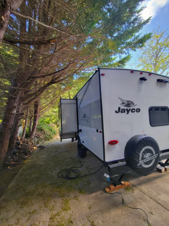 A white Jayco travel trailer, a type of RV, with a slide-out extended, parked among trees and logs. Great RV rental option!. Jayco Jay Flight SLX 197MBW Baja Edition 2025