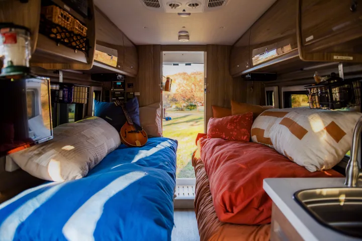 Bright RV rental interior: two cozy beds, one with a mandolin, overhead storage, and a sink, overlooking a scenic outdoor view.. Winnebago Travato 2020