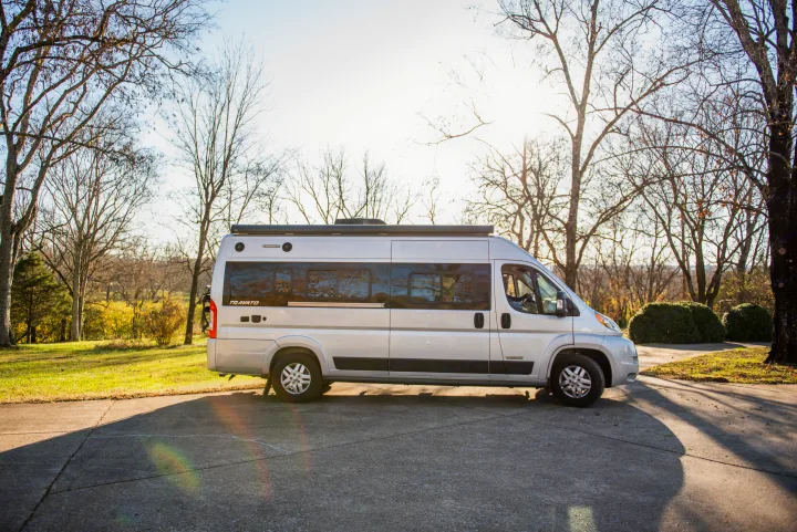 Silver Winnebago Travato camper van rv rental, parked on a sunlit pavement amidst bare trees and green grass.. Winnebago Travato 2020
