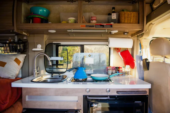 Well-equipped rv rental kitchen in a camper van, showing a sink, stove, and organized overhead storage for cooking supplies.. Winnebago Travato 2020