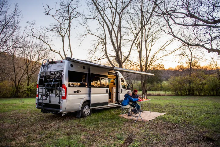 An RV rental camper van parked in a grassy field at sunset, with two people enjoying dinner outside under an awning.. Winnebago Travato 2020